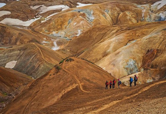 Eine Wandergruppe beschreitet einen schmalen Pfad in den karg-braunen Rhyolithbergen des isländischen Hochlands.