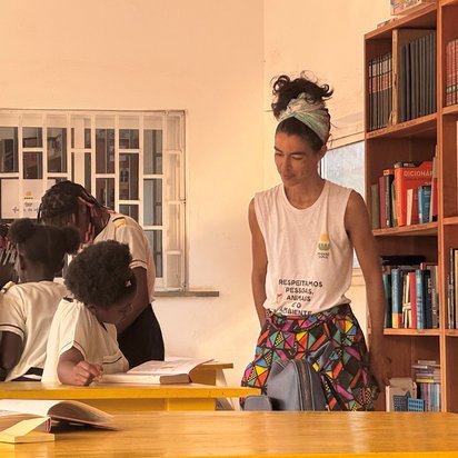Links im Bild sitzt ein afrikanisches Mädchen und liest in einem Buch. Rechts neben ihr steht eine Frau und beobachtet sie dabei. Die rechte Wand wird von einem Bücherregal eingenommen.