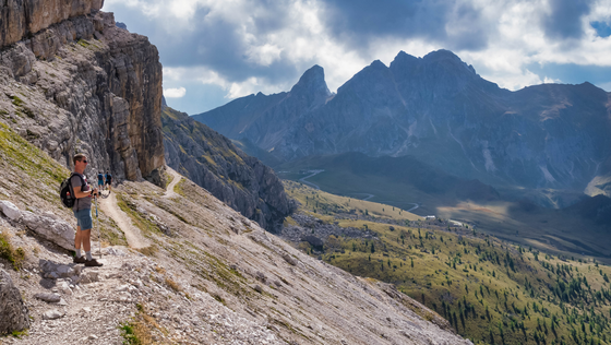 Wanderung im Val di Fassa Vorne links an einem Berghang steht ein Wanderer, im Hintergrund ragen die Dolomiten empor.