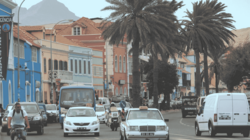 Eine vielbefahrene palmengesäumte Straße führt entlang einer bunten Häuserzeile. Hinter den Häusern erkennt man Berge.