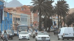 Eine vielbefahrene palmengesäumte Straße führt entlang einer bunten Häuserzeile. Hinter den Häusern erkennt man Berge.