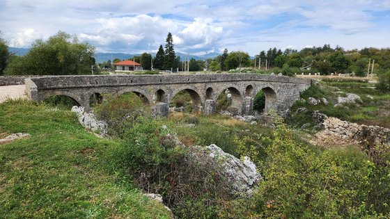 Über einen grün-bewucherten ausgetrockneten Flusslauf erstreckt sich eine aus Steinquadern errichtete, antike Brücke.