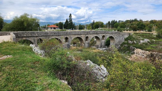 Über einen grün-bewucherten ausgetrockneten Flusslauf erstreckt sich eine aus Steinquadern errichtete, antike Brücke.