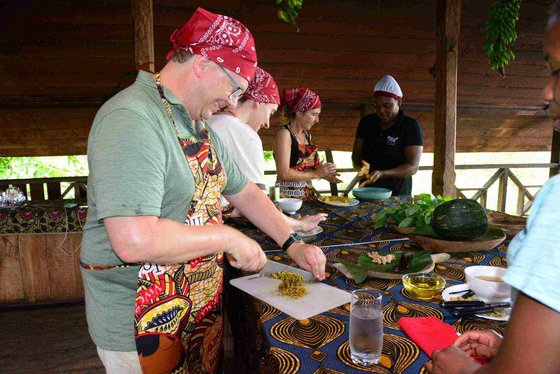An einem mit einer bunten Tischdecke belegten Tisch stehen mehrere weiße Menschen in Kochschürzen und roten Kopftüchern und bereiten angeleitet von zwei schwarzen Frauen Speisen zu.