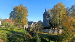 Rechts im Bildmittelgrund erhebt sich eine aus Holz gebaute, von einer Steinmauer umgebene Kirche. Links von ihr stehen nebeneinander ein paar Häuser. Im Vordergrund sieht man eine begrünte Landschaft mit Wiese und Bäumen. Der Himmel ist strahlend blau.