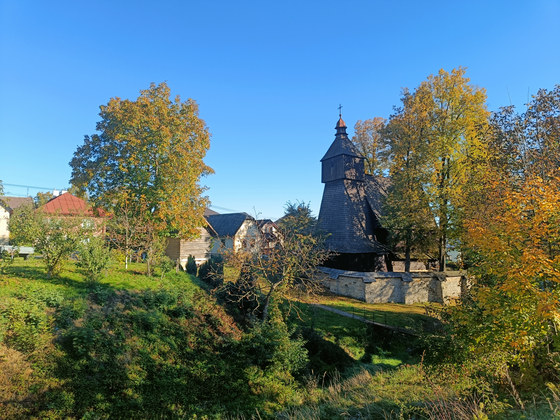Rechts im Bildmittelgrund erhebt sich eine aus Holz gebaute, von einer Steinmauer umgebene Kirche. Links von ihr stehen nebeneinander ein paar Häuser. Im Vordergrund sieht man eine begrünte Landschaft mit Wiese und Bäumen. Der Himmel ist strahlend blau.