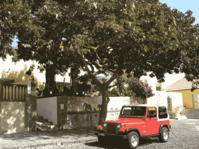 Ein roter Jeep steht auf einer Straße unter einem Baum. Auf der höher gelegenen Terrasse dahinter sitzen mehrere Männer.