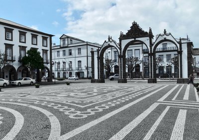 Das Stadttor, Wahrzeichen Ponta Delgadas Auf einem Platz mit hell- und dunkelgrauem Kopfsteinpflaster steht ein dreibogiges Tor. Den Platz umgeben historische Gebäude.