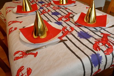 Eine Festtafel ist mit einer Tischdecke mit Krebsmotiven, roten Servietten und goldenen Hütchen geschmückt.