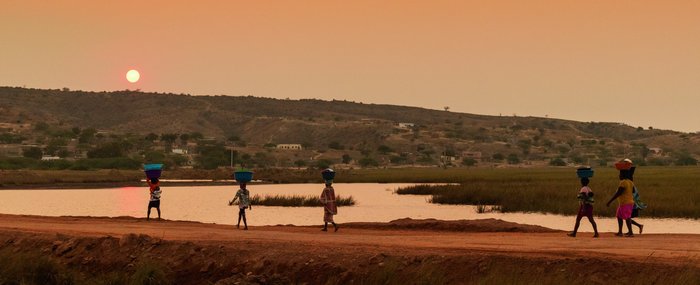 Einheimische Frauen laufen bei Sonnenuntergang mit Wannen auf dem Kopf eine Straße an einem Gewässer entlang