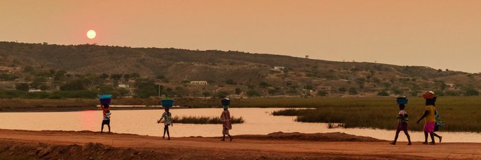 Einheimische Frauen laufen bei Sonnenuntergang mit Wannen auf dem Kopf eine Straße an einem Gewässer entlang