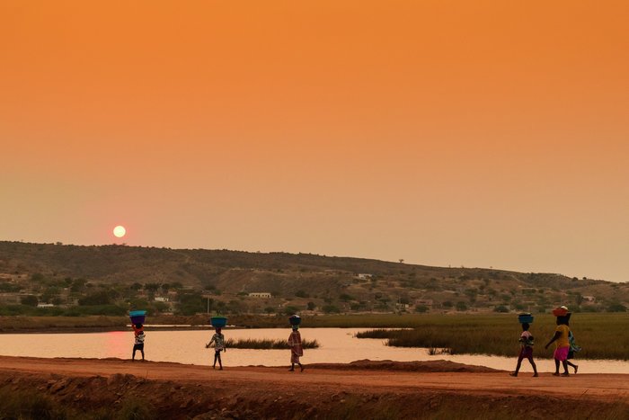 Einheimische Frauen laufen bei Sonnenuntergang mit Wannen auf dem Kopf eine Straße an einem Gewässer entlang