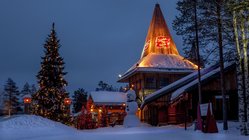 Ein mit Lichterketten geschmücktes Haus steht flankiert von einem Weihnachtsbaum in einer Schneelandschaft.