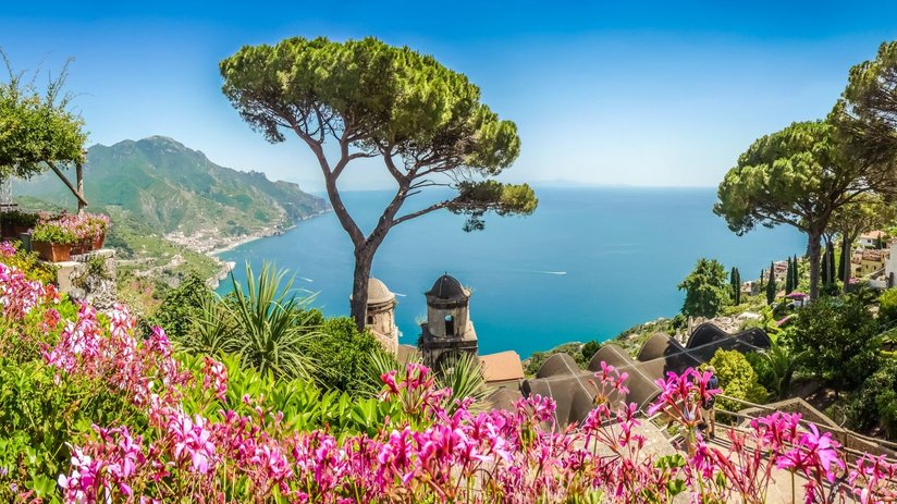 Von einer Erhebung schaut man über blühende Blumen und durch Zypressen auf den Golf von Salerno.