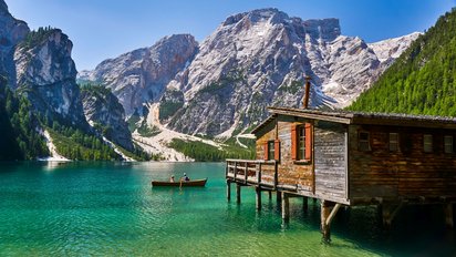 Die untere linke Bildhälfte wird von einer grünen Wasserfläche eingenommen, auf der zwei Ruderboote unterwegs sind. Rechts im Vordergrund befindet sich eine Holzhütte, die auf Stelzen im Wasser steht. Hinter der Wasserfläche erheben sich karge, steile Berge, die in den tieferen Lagen von Nadelbäumen bewachsen sind.