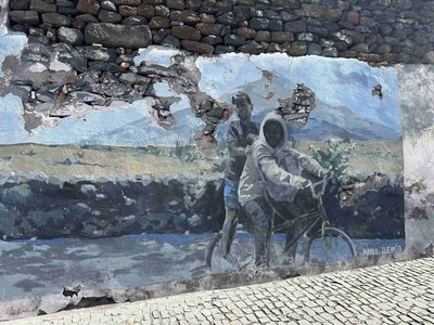 Die Wandgemälde in  São Filipe auf Fogo gehören zu den Sehenswürdigkeiten der Kapverden. Dieses zeigt zwei afrikanische Jungs in Streetwear auf einem Fahrrad, im Hintergrund eine gebirgige kapverdische Landschaft. Das Bild ist schon recht verwittert und man kann auf die Mauer aus großen Natursteinen durchblicken.