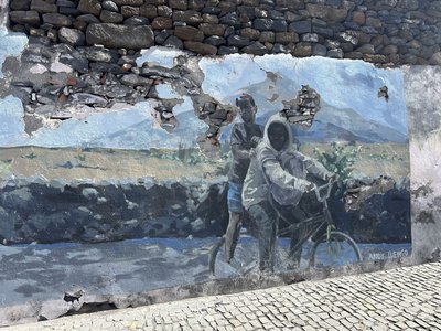 Die Wandgemälde in  São Filipe auf Fogo gehören zu den Sehenswürdigkeiten der Kapverden. Dieses zeigt zwei afrikanische Jungs in Streetwear auf einem Fahrrad, im Hintergrund eine gebirgige kapverdische Landschaft. Das Bild ist schon recht verwittert und man kann auf die Mauer aus großen Natursteinen durchblicken.