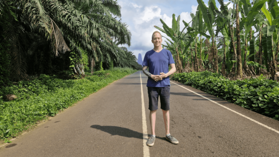Ein junger Mann steht auf einer kleinen, zweispurigen Straße, die von tropischer Vegetation umgeben ist.