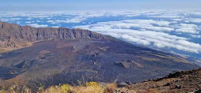 Blick vom Pico do Fogo Man schaut von einer vulkanischen Erhebung hinab über Hänge mit schwarzer Lavaasche bis hinunter zum Meer, das jedoch von Wolken überfangen ist.