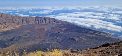 Man schaut von einer vulkanischen Erhebung hinab über Hänge mit schwarzer Lavaasche bis hinunter zum Meer, das jedoch von Wolken überfangen ist.