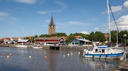 Man blickt vom Wasser aus auf einen kleinen Hafen, in dem drei kleine Segelboote liegen. Entlang des Ufers stehen mehrere niedrige Gebäude, dahinter Bäume, aus denen sich ein Kirschturm erhebt. Die obere Bildhälfte wird von blauem Himmel mit wenigen weißen Wolken eingenommen.