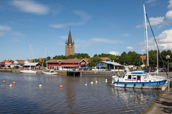 Man blickt vom Wasser aus auf einen kleinen Hafen, in dem drei kleine Segelboote liegen. Entlang des Ufers stehen mehrere niedrige Gebäude, dahinter Bäume, aus denen sich ein Kirschturm erhebt. Die obere Bildhälfte wird von blauem Himmel mit wenigen weißen Wolken eingenommen.