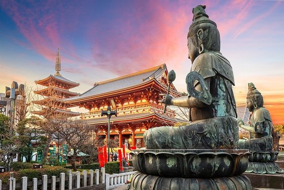 Eine seitlich fotografierte Buddha-Statue. Im Hintergrund befindet sich der Sensoji-Tempel unter dem rosa-bläulichen Himmel.