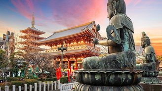 Eine seitlich fotografierte Buddha-Statue. Im Hintergrund befindet sich der Sensoji-Tempel unter dem rosa-bläulichen Himmel.