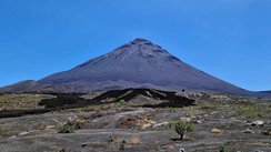 Der Vordergrund wird von einer schwarzen Vulkanaschenlandschaft ausgefüllt. Im Hintergrund erhebt sich ein spitzer Vulkankegel umfangen von leuchtend blauem Himmel.