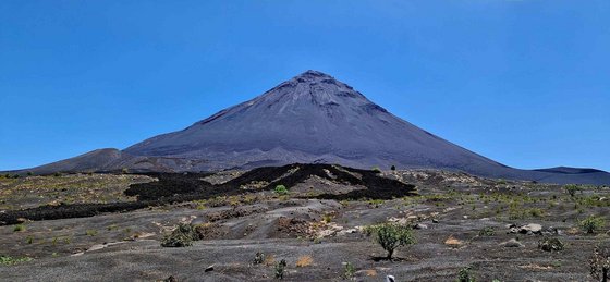 Der Vordergrund wird von einer schwarzen Vulkanaschenlandschaft ausgefüllt. Im Hintergrund erhebt sich ein spitzer Vulkankegel umfangen von leuchtend blauem Himmel.