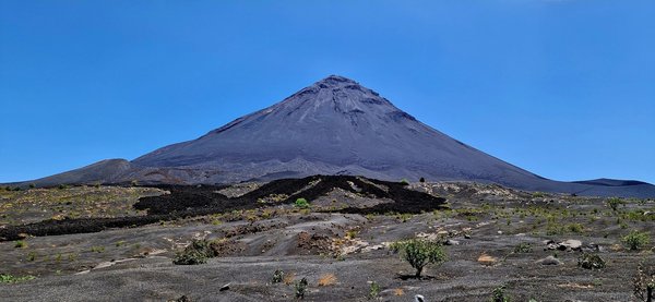 Der Vulkan Pico do Fogo Der Vordergrund wird von einer schwarzen Vulkanaschenlandschaft ausgefüllt. Im Hintergrund erhebt sich ein spitzer Vulkankegel umfangen von leuchtend blauem Himmel.