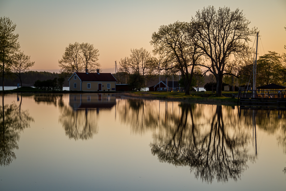 Sjötorp am westlichen Ende des Göta-Kanals Die untere Bildhälfte wird von einer spiegelnden Wasserfläche eingenommen, die die dahinterliegenden Bäume sowie ein gelbes Haus reflektiert.