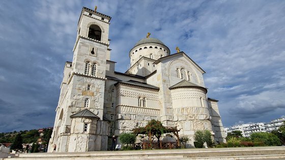 Das Foto zeigt aus der Froschperspektive eine orthodoxe Kirche von der Seite, so dass die Fassade links im Bild ist. Links erhebt sich einer der die Fassade flankierenden Türme, in der Mitte die Kirchenkuppel. Die Kirche ist weiß mit grauen Dächern und von mehreren goldenen Kreuzen gekrönt. Sie ist von einem blau-grauen Himmel umfangen.