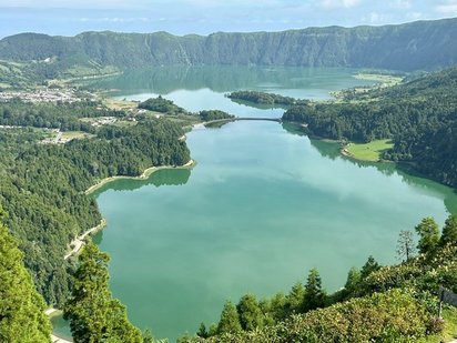 Das Foto wird von einer grünen Wasserfläche, die einer unregelmäßig geformten liegenden Acht gleicht, eingenommen. Der See ist auf allen Seiten von bewaldeten Hängen umgeben.