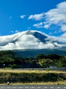Das hochformatige Bild wird in der unteren Hälfte von grüner Landschaft eingenommen. In der Mitte erhebt sich ein von Wolken umhüllter Vulkankegel, darüber im oberen Bilddrittel der blaue Himmel, auf dem auf der rechten Seite ebenfalls weiße Wolken treiben.