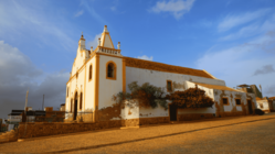 An einer leicht ansteigenden Kopfstein-gepflasterten Straße liegt eine weißgetünchte Kirche, deren Fassade mit gelben Schmuckelementen versehen ist. Darüber blauer Himmel