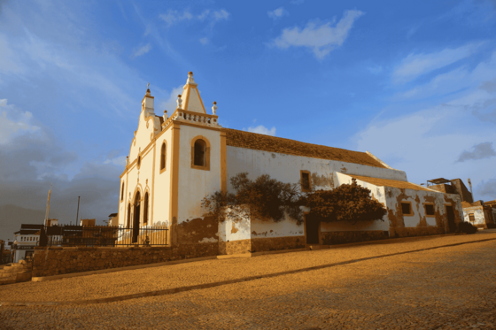 An einer leicht ansteigenden Kopfstein-gepflasterten Straße liegt eine weißgetünchte Kirche, deren Fassade mit gelben Schmuckelementen versehen ist. Darüber blauer Himmel
