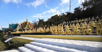 Der Wat Phou bei Paksé Im Vordergrund stehen viele goldene Buddha-Statuen in Reihen nebeneinander. Links im Hintergrund erheben sich mehrere kleinere Tempel.