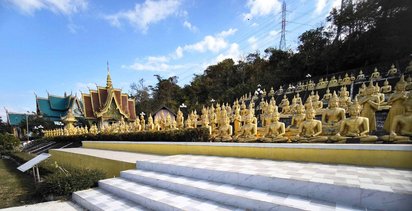 Im Vordergrund stehen viele goldene Buddha-Statuen in Reihen nebeneinander. Links im Hintergrund erheben sich mehrere kleinere Tempel.