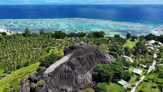 Luftaufnahme von einem massiven Granitfelsen, der inmitten üppiger Vegetation und Palmen, mit Blick auf das blaue Meer liegt.