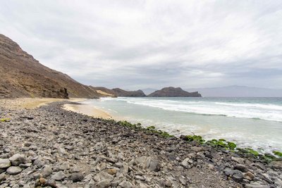 Der ursprüngliche Küstenort Salamansa auf  São Vicente und die Wandermöglichkeiten in seiner Umgebung gehören zu den Top-Sehenswürdigkeiten der Kapverden. Das Foto zeigt eine menschenleere weite Kiesstrandlandschaft mit dem Meer zur Rechten und sich im Mittel- und Hintergrund daraus erhebenden Hügeln. Das gesamte Bild wird von Grautönen dominiert.
