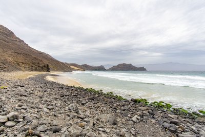 Der ursprüngliche Küstenort Salamansa auf  São Vicente und die Wandermöglichkeiten in seiner Umgebung gehören zu den Top-Sehenswürdigkeiten der Kapverden. Das Foto zeigt eine menschenleere weite Kiesstrandlandschaft mit dem Meer zur Rechten und sich im Mittel- und Hintergrund daraus erhebenden Hügeln. Das gesamte Bild wird von Grautönen dominiert.