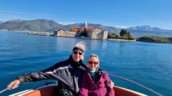 Ein mit Windjacken bekleidetes Paar sitzt im Bug eines kleinen Bootes. Über die blaue Wasserfläche sieht man auf eine Stadt am Ufer. Die Landschaft dahinter besteht aus niedrigen Hügeln.