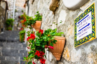 An einer Hausmauer hängt ein Schild zum Wanderweg "Weg der Götter", dahinter hängen Blumenkästen mit rotblühenden Blumen.