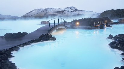 Die Blaue Lagune auf Island Das Bild füllt ein dampfendes Naturschwimmbecken aus, das von einem Weg und einer Brücke überspannt wird. Im Hintergrund sieht man einen schneebedeckten Hügel.