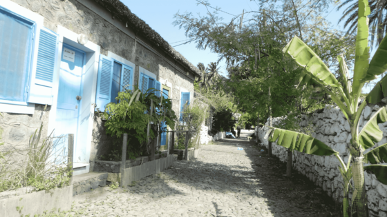 Man sieht eine mit Kopfsteinpflaster belegte Straße, die auf der rechten Seite von einer Bruchsteinmauer eingefasst ist, links der Straße erstreckt sich eine Häuserzeile ebenfalls aus groben Steinen erbaut.