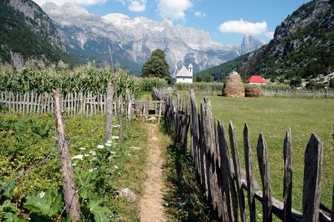 Ein Holzzaun säumt den Weg zu einem kleinen Bauernhof vor einem beiindruckenden Alpenpanorama