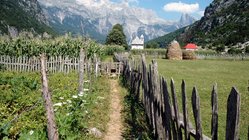 Ein Holzzaun säumt den Weg zu einem kleinen Bauernhof vor einem beiindruckenden Alpenpanorama