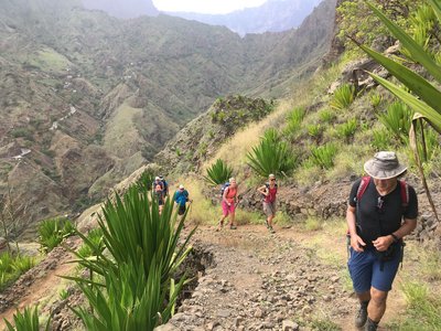 Wandern in der Serra Malagueta Der Betrachter sieht eine Gruppe Wanderer bergan auf sich zukommen. Die umgebenden Hänge der Serra Malagueta auf den Kapverden sind mit Agaven bewachsen. Die links hintere Bilddiagonale wird von einem Tal mit steil aufragenden Bergflanken eingenommen. Das Bild wird von Grüntönen dominiert.