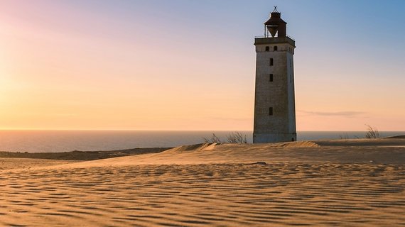 Ein steinerner Leuchtturm im rosa-orangefarbenen Licht der Dämmerung am Strand.